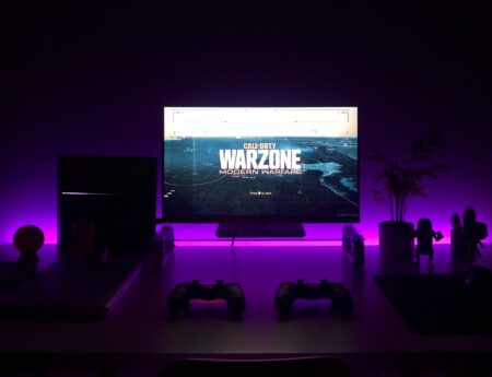 A gaming setup featuring a monitor displaying "Call of Duty Warzone: Modern Warfare," illuminated by purple backlighting. The desk holds two controllers, speakers, and various figurines, creating a lit ambiance