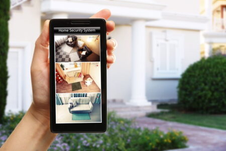 A person's hand holding a smartphone displaying a home security app with a live camera feed of the house interior, set against the backdrop of the front yard.