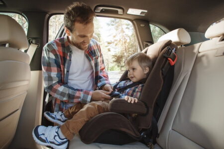 A father smiles at his young son while fastening him into a car seat in a vehicle, surrounded by a secure forested area in daylight.