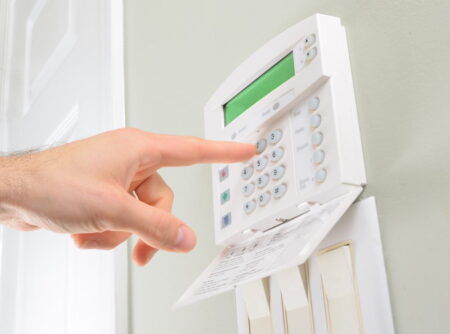 A person's hand pressing a button on a white wall-mounted home security alarm keypad with a green display screen.