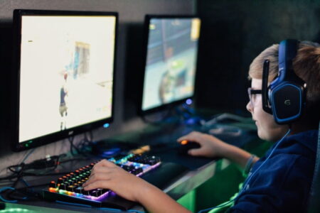 A young boy wearing headphones is focused on playing a video game on a computer, with colorful keyboard lights visible in a dimly lit room. He hopes to make money through competitive gaming.