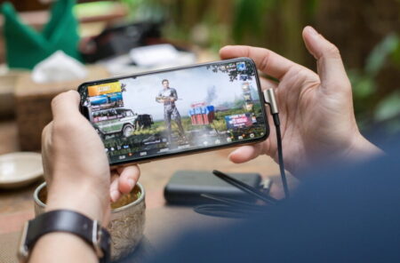 A person's hands holding a smartphone displaying an action game with cars and a character on the screen, at a table with greenery in the background, showing how games make money.
