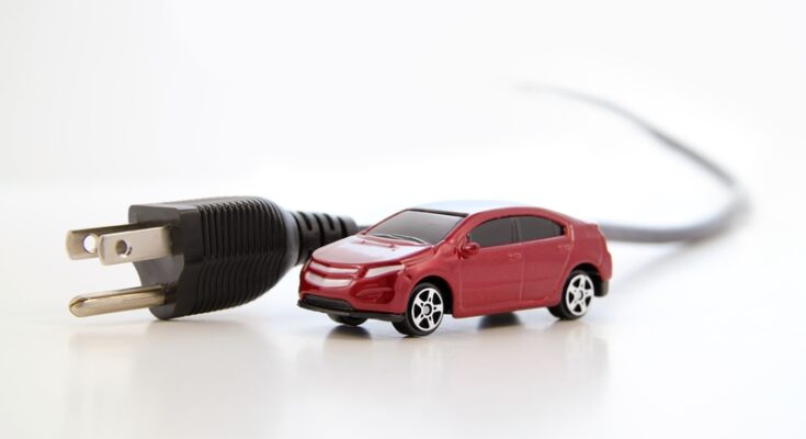 A small red electric car connected to a black electrical plug with a cord, symbolizing electric vehicles, set against a clean, white background.