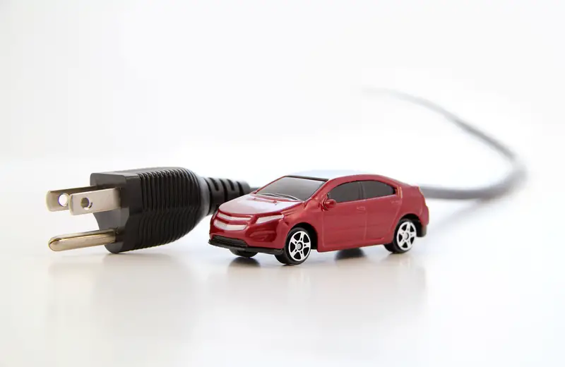 A small red electric car connected to a black electrical plug with a cord, symbolizing electric vehicles, set against a clean, white background.