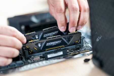 A person performs a PC upgrade by installing RAM modules branded Ballistix into a computer motherboard, carefully aligning the stick correctly in the memory slot.