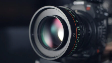 Close-up of a video camera lens focusing on 85mm, with reflections of light on the glass and visible aperture and distance scale markings.
