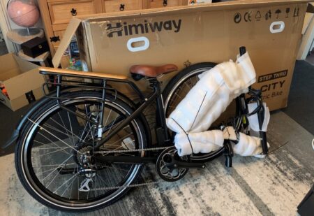 A new black Himiway City Pedelec electric bike, partially covered with protective foam and plastic, stands next to its open cardboard shipping box in a room.