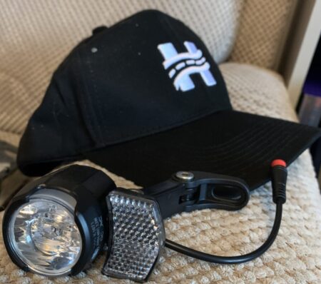A black baseball cap with a white logo on it, next to a black Himiway City Pedelec bike light with multiple LEDs, placed on a beige textured surface.