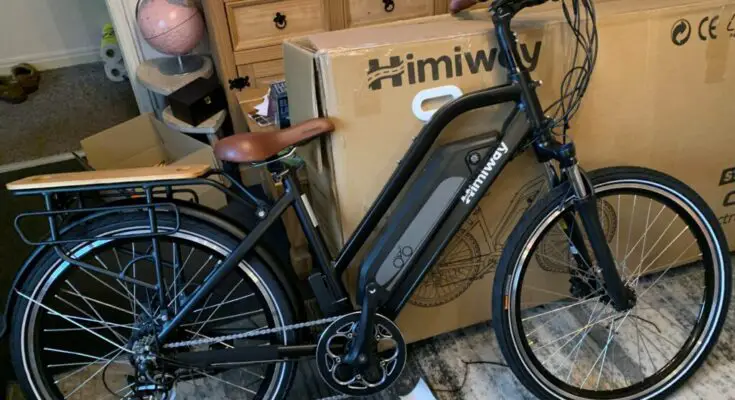 A Himiway City Pedelec with a black frame and brown saddle stands in front of cardboard boxes in a room. The eBike features pedal assist, a rear rack, and a head