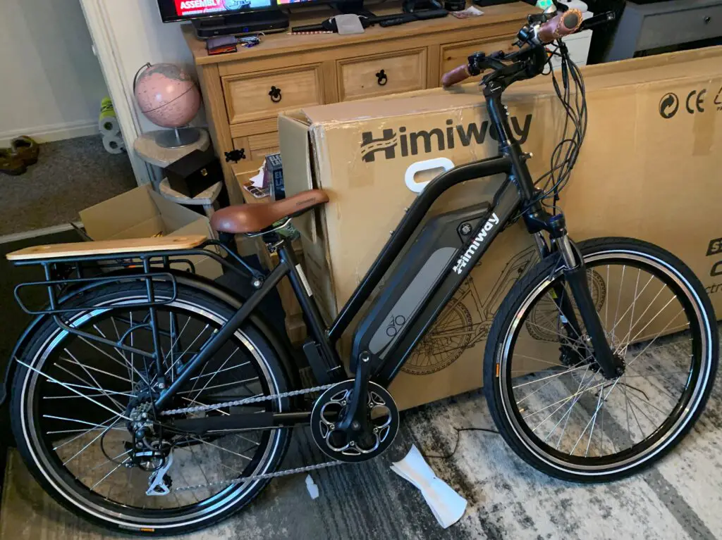 A Himiway City Pedelec with a black frame and brown saddle stands in front of cardboard boxes in a room. The eBike features pedal assist, a rear rack, and a head
