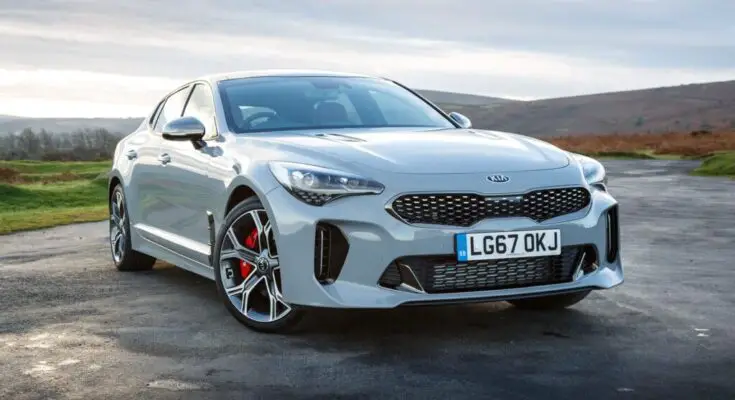 A silver Kia Stinger car parked on an open road with a scenic, hilly landscape in the background during the daytime.