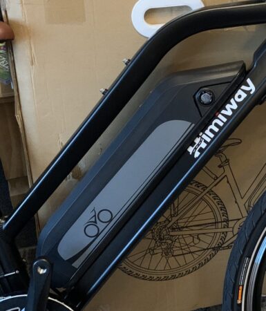 Close-up of a Himiway City Pedelec frame, showcasing the battery labeled "himiway" with a bicycle icon, against a blurred background featuring cardboard boxes.
