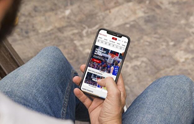 A person sitting and browsing a sportsbooks website on a smartphone held in their hands, focusing on a basketball game article.