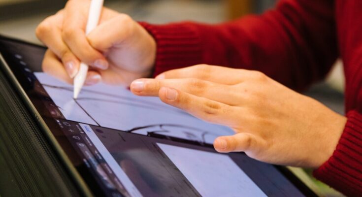 A person in a red sweater using a stylus on a digital drawing tablet, focusing on a creative design project with new technologies for students. Their fingers are visible on the screen as they work.