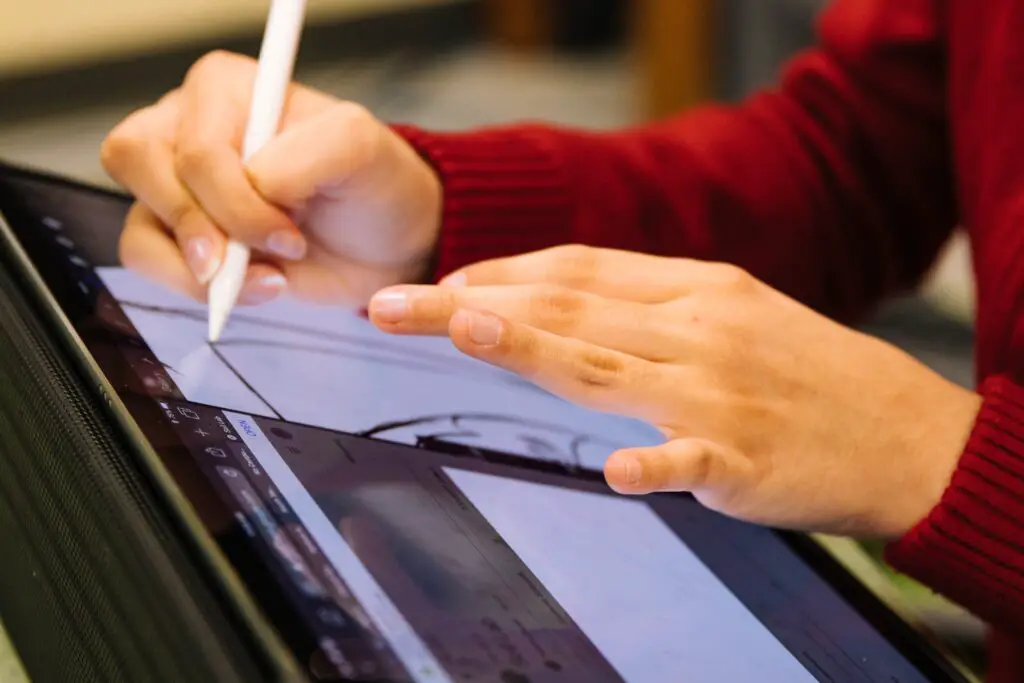 A person in a red sweater using a stylus on a digital drawing tablet, focusing on a creative design project with new technologies for students. Their fingers are visible on the screen as they work.
