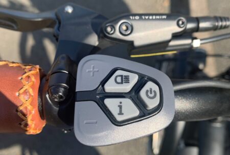 Close-up of a Himiway City Pedelec's handlebar, showing a control pad with buttons for power and info, plus "+" and "-" for adjustments, next to a brown leather grip.