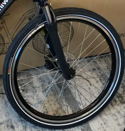 A close-up of a Himiway City Pedelec bicycle wheel with a black tire, detailed spokes, and fork visible, set against a cardboard background with part of another wheel in view.