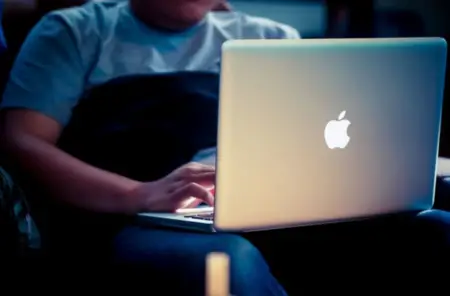 Person sitting in a dimly lit room, using a Mac on their lap, focused on the glowing screen. Only their hands and part of the laptop are visible.