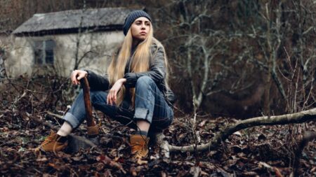 A young woman with long blonde hair, wearing a gray beanie, denim jeans, and a camouflage jacket, sits pensively on a fallen tree branch in a wooded area with a small cabin in the
