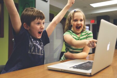 Two excited children, a boy and a girl, are sitting at a desk and raising their arms in joy while looking at the latest video game trends on a laptop screen. The boy is wearing a grey