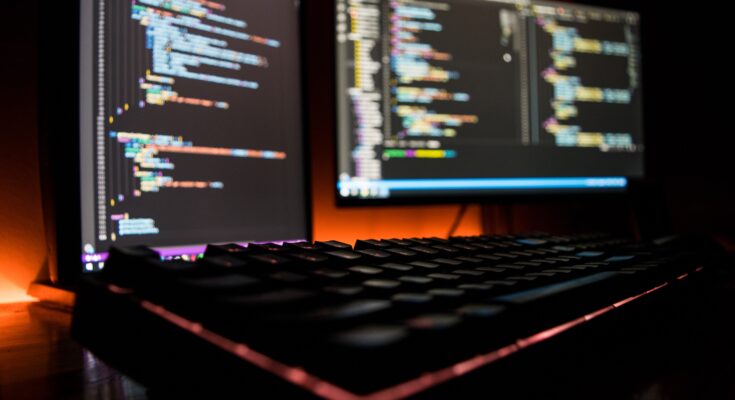 Two computer screens displaying colorful poker software platforms in a dark room, with a blurred mechanical keyboard in the foreground illuminated by ambient light.