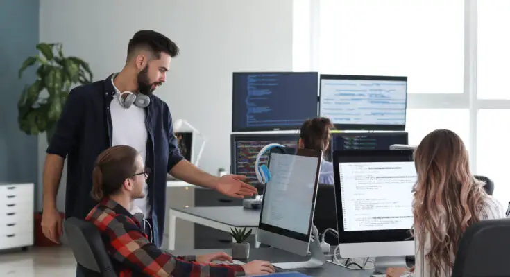 A group of four colleagues, including three men and one woman, engaged in discussion and work at a tech office with multiple computer screens displaying code, offering enhanced IT support.