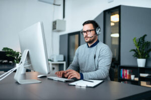 A focused IT Support specialist wearing headphones and glasses is working at his desk, typing on a keyboard with a large monitor, in a modern office setting.