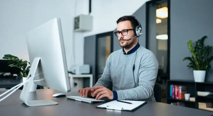 A focused IT Support specialist wearing headphones and glasses is working at his desk, typing on a keyboard with a large monitor, in a modern office setting.