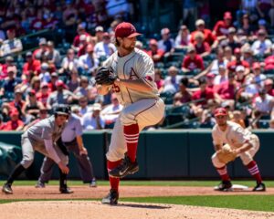 A baseball pitcher, one of the oddest sports stars, in a white and red uniform mid-pitch during a sunny game, with spectators in the background and a catcher and umpire in position.