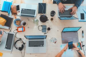 An overhead view of a busy digital workplace with four people using laptops, surrounded by notepads, snacks, smartphones, and coffee mugs on a wooden table.