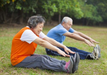 An elderly Asian couple happily exercising in a park, doing stretching exercises on the grass despite the poor air quality. The woman smiles while reaching towards her toes; the man, in blue, also stretches.