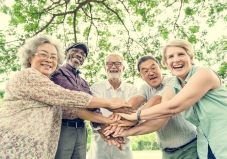A diverse group of joyful seniors with hands stacked together in a park, smiling broadly, symbolizing teamwork and friendship under a large tree, enjoying the fresh air quality.