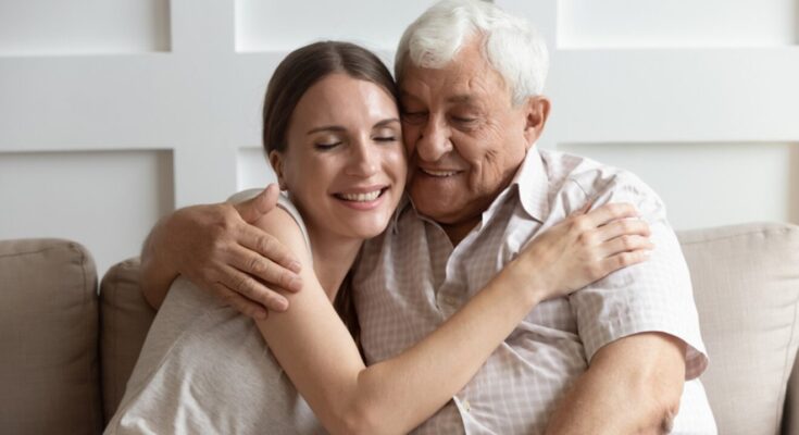 A young woman and an elderly man embrace warmly, smiling as they sit closely on a sofa in a neutrally decorated room with optimal air quality, expressing love and comfort.