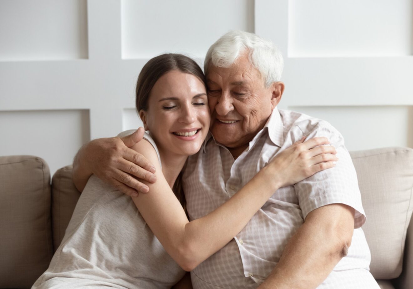 A young woman and an elderly man embrace warmly, smiling as they sit closely on a sofa in a neutrally decorated room with optimal air quality, expressing love and comfort.