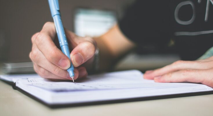 person holding blue ballpoint pen writing in notebook