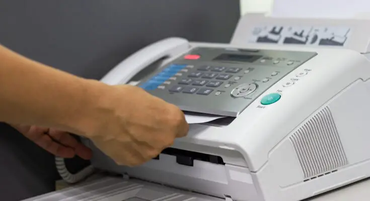 A person's hand is removing a sheet of paper from a multifunction printer with Internet Faxing capabilities in an office setting. The printer has several control buttons and a paper output tray.