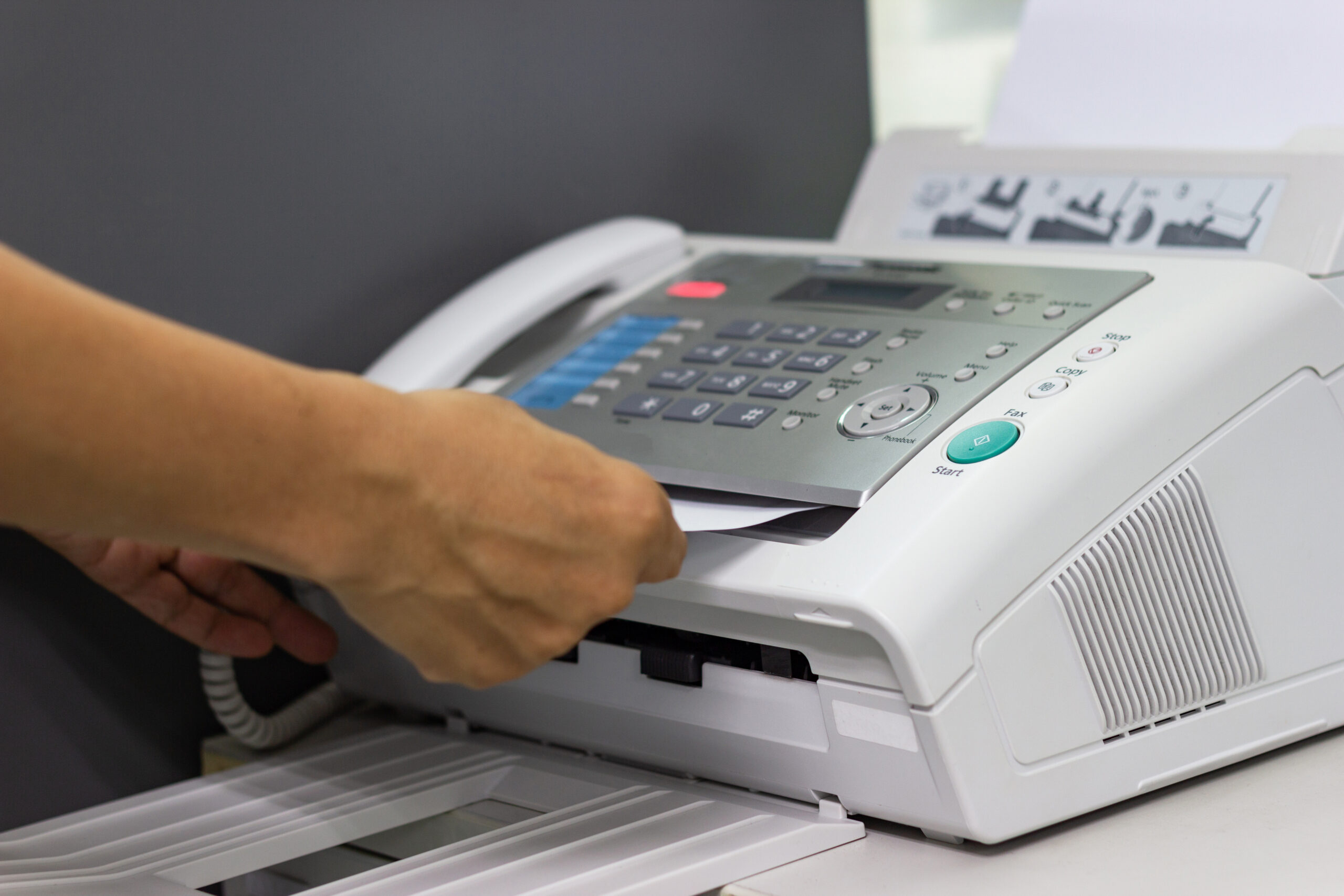 A person's hand is removing a sheet of paper from a multifunction printer with Internet Faxing capabilities in an office setting. The printer has several control buttons and a paper output tray.