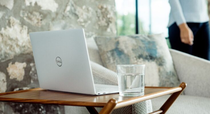 A dell laptop worth sits on a wooden tray table beside a glass of water, with a gray sofa and partial view of a standing person in the background, against a stone wall.