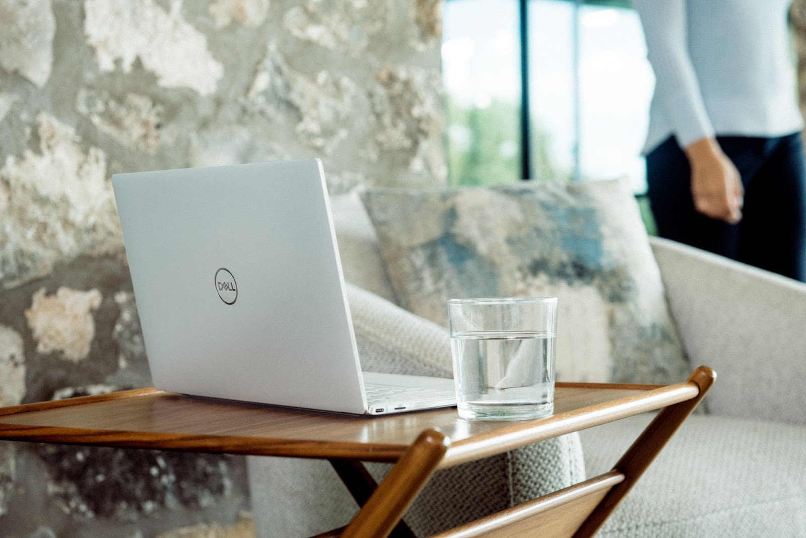 A dell laptop worth sits on a wooden tray table beside a glass of water, with a gray sofa and partial view of a standing person in the background, against a stone wall.