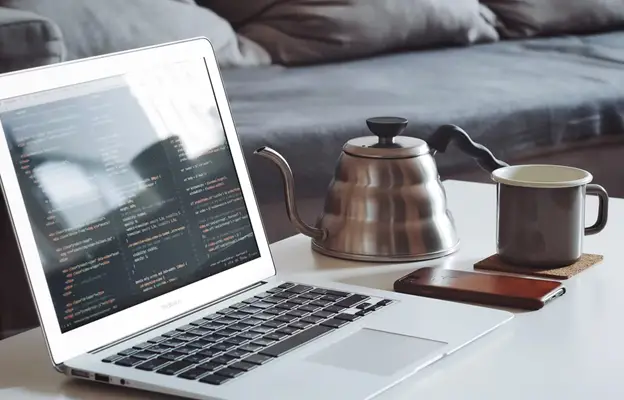 A cozy workspace featuring an open laptop displaying code, accompanied by a teapot, a mug, and a smartphone with the fastest VPN on a table near a couch.