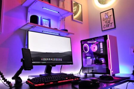 A vibrant gaming setup featuring a gaming desktop with rgb lighting, dual monitors, a backlit mechanical keyboard, and a gaming mouse, all in a room with colorful ambient lighting and motivational posters.