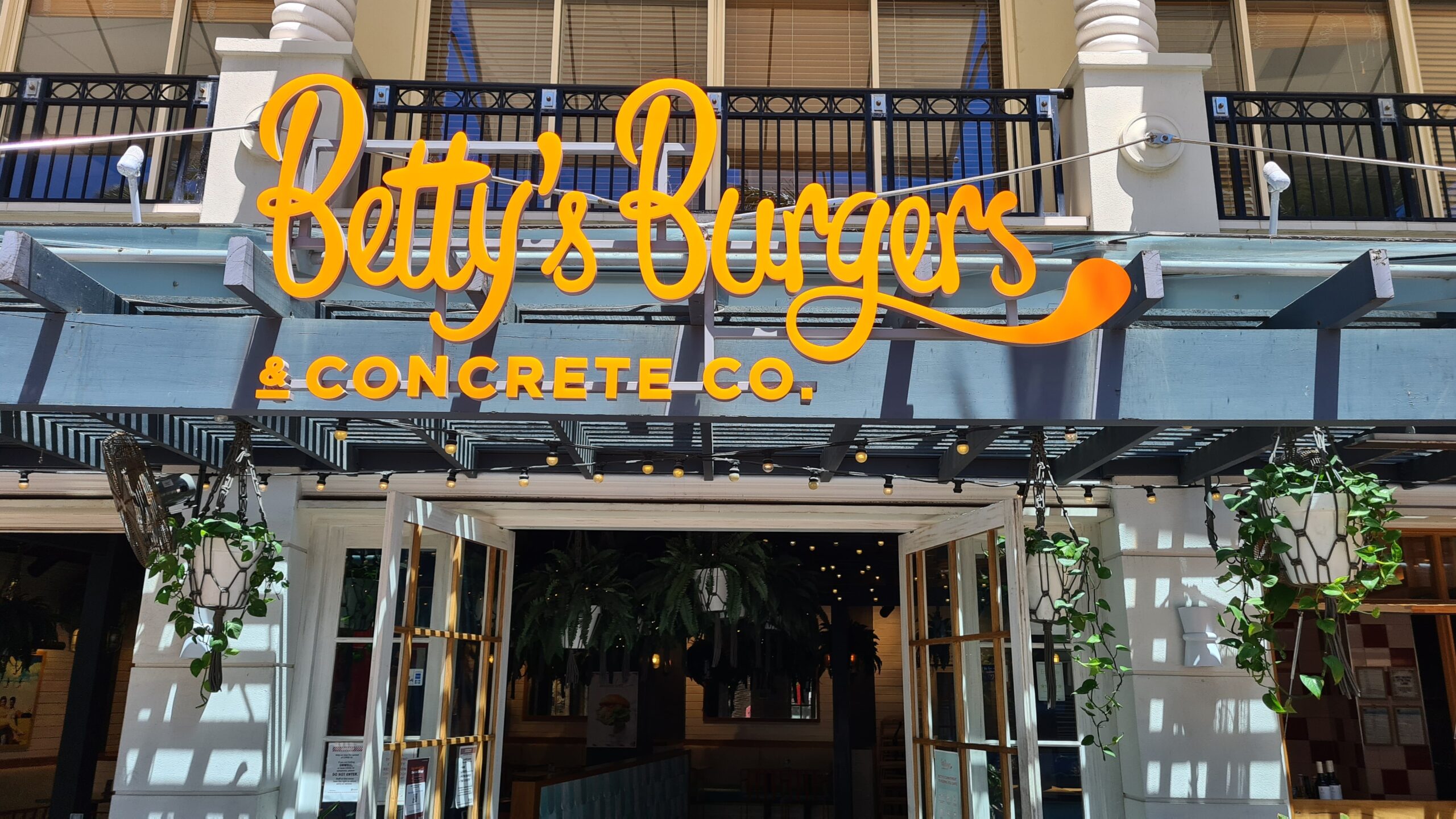 Bright neon sign displaying "betty's burgers & concrete co." co-branding hung on a blue storefront with hanging green plants and an open entrance.