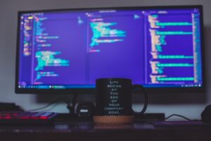 A desk with a coffee mug featuring an inspirational quote in front of a computer screen displaying web applications security code, illuminated in soft blue light.