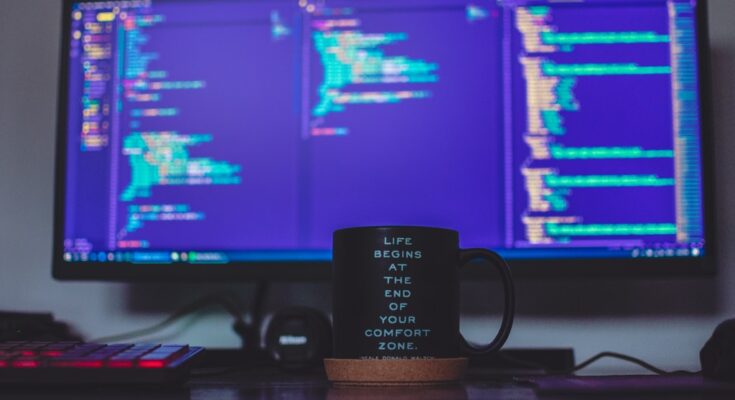 A desk with a coffee mug featuring an inspirational quote in front of a computer screen displaying web applications security code, illuminated in soft blue light.