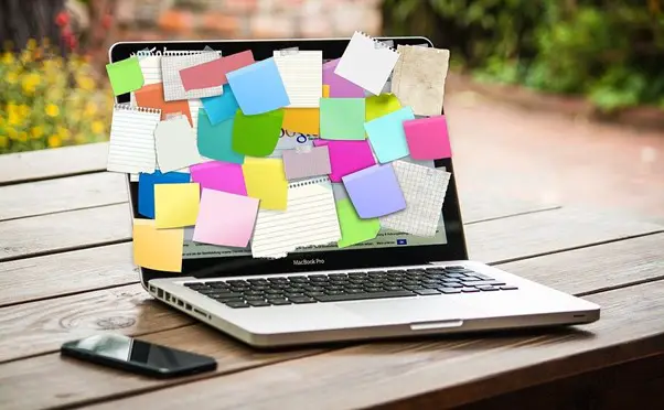 An open laptop on a wooden table covered with colorful *online sticky notes* and scraps of paper on its screen, alongside a smartphone.