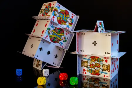 A house of cards intricately built in a multi-level design, with various playing cards displaying kings, queens, and other figures typically found in casinos online, alongside colorful dice scattered around on a dark background