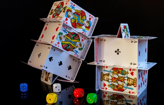 A house of cards intricately built in a multi-level design, with various playing cards displaying kings, queens, and other figures typically found in casinos online, alongside colorful dice scattered around on a dark background