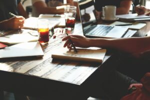 A group of people collaborating around a wooden table with laptops, notebooks, pens, and drinks illuminated by natural sunlight are discussing how to provide their customers with the best service possible.