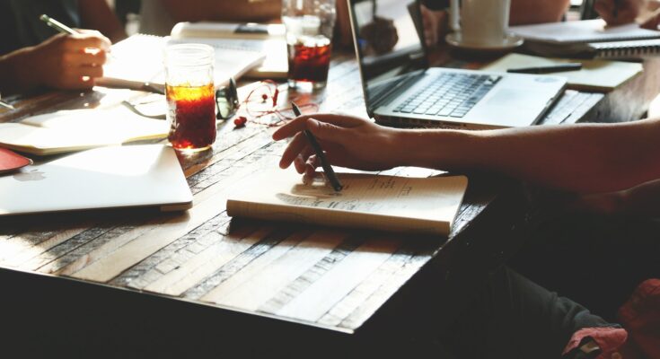 A group of people collaborating around a wooden table with laptops, notebooks, pens, and drinks illuminated by natural sunlight are discussing how to provide their customers with the best service possible.