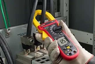 A person wearing gloves uses a clamp meter to measure electrical voltage in an industrial setting.